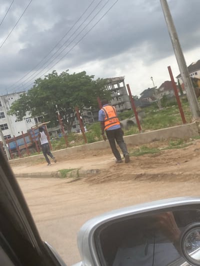 The scene is in Iddo Tudunwada, Nigeria, viewed from inside a vehicle, with a side mirror visible. A dirt road runs parallel to an unfinished construction site delineated by red steel posts. Two men walk on the road. One wears an orange reflective vest over a blue shirt and dark trousers, while the other wears a white shirt and dark trousers. A building with white walls and grid windows is in the background. The sky is overcast.