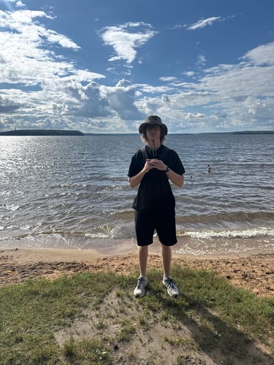 A person stands on a sandy beach next to a body of water. The person is wearing a black t-shirt, black shorts, a bucket hat, and athletic shoes. They have their hands clasped in front of them and are looking towards the camera. The water is wavy and reflects the sunlight. Behind the water, a treeline is visible in the distance, suggesting a natural, possibly rural setting. The sky is blue with scattered white clouds, indicating fair weather. The scene appears to be outdoors during the daytime. The overall mood is casual and relaxed, with the person appearing to be enjoying a day by the water. The image appears to be a candid shot, possibly taken by a friend or companion.