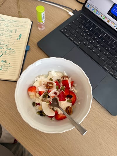 A top-down, slightly oblique view displays a workspace on a light-colored wooden or laminate tabletop, located in the city of Anavissos Municipal Unit, Greece. In the foreground, a white, scallop-edged ceramic bowl contains a meal consisting of sliced strawberries, large coconut flakes, whole pecans, various seeds (including pumpkin and possibly sunflower), and what appears to be a creamy base such as yogurt or cottage cheese. A silver spoon with a square tip rests within the bowl.

To the left of the bowl, an open spiral-bound notebook with yellowed pages and a blue spine contains handwritten notes in green and black ink. Visible text includes phrases like "Ancient Messini," "Nestor's Palace," "tasting + food pairing," "kids come along?", and numerical entries such as "2 days," "30-40 photos," "5-6 clips," "5/5," "hotel," and "2-3 days."

Between the notebook and a laptop, a small, translucent bottle with a neon green cap stands upright. It contains a clear liquid with a slight pinkish hue.

In the upper right portion of the frame, a dark gray laptop, consistent with a MacBook Pro, is open with its screen displaying multiple application windows, including what appears to be a web browser with several tabs open. A white USB-C cable is connected to the left side of the laptop, extending upwards and out of frame. The black keyboard and trackpad are visible. No individuals are present in the scene.