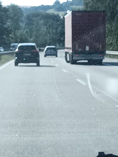 The image shows a view from a vehicle traveling on a highway, likely in Šternov, Czechia. The foreground is the blurred asphalt of the road, with some white lines visible. In the middle ground, several vehicles are driving on the highway. Closest to the viewer is a dark-colored car, followed by a white car and then a dark-colored car. To the right, a large red and white truck is also traveling on the highway. The background features a dense line of trees and hills under a bright, overcast sky, suggesting it is daytime. The overall impression is a typical highway scene with moderate traffic on a clear day. The image is taken through a window, indicated by some smudges and streaks on the glass. No text is visible in the image.