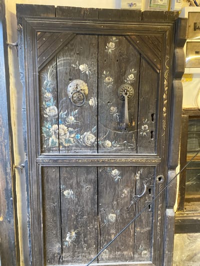 The image shows a close-up view of an antique, dark-stained wooden chest or cabinet door. The door is decorated with painted floral designs, primarily white and blue roses. The wooden planks are heavily textured and weathered, indicating age. The door is divided into two sections, with a decorative vertical strip separating them. Each section has metal hardware, including hinges and handles. The surrounding frame is also made of dark wood and has additional decorative elements. The context of the scene suggests this item is located in Konya, Turkey, given the style and origin of the object.