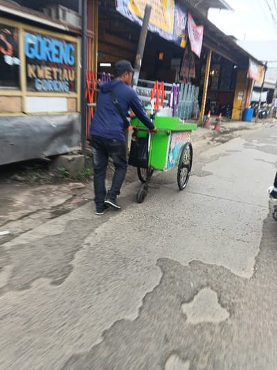 This image depicts a street scene in Margahayu, Indonesia, under daylight conditions. The primary subject is an adult male, viewed from behind, wearing a dark blue long-sleeved top, dark jeans, dark shoes, and a dark baseball cap. He has a black shoulder bag slung over his left shoulder. He is actively pushing a three-wheeled vending cart, predominantly bright green, with two large spoked wheels at the rear and a smaller caster wheel at the front. The cart has faded graphics on its side. A black bag is hanging from the left side of the cart.
To the left of the man and cart, a light-colored building features a sign with blue lettering that reads "GORENG" and "KWETIAU GORENG," indicating a fried noodle establishment. The building has a dark-colored base. To the right, a series of open-fronted stalls or shops with sloped corrugated roofs are visible in the background. These stalls display various items, including stacks of purple and orange plastic chairs or stools. Colorful banners and flags are draped across the front of these structures. Further in the distance, a blue barrel and orange traffic cones are discernible.
The ground is a paved surface, likely asphalt or concrete, exhibiting several dark, irregular wet patches, suggesting recent moisture. A partial view of a foot wearing a dark shoe with a white sole is visible in the lower right corner of the frame, indicating the presence of another individual outside the main focus.