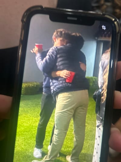 A photo of an iPhone displaying a photograph of two people standing on artificial turf. One person is wearing a dark puffer jacket and khaki pants, holding a red cup. The other person is wearing a dark puffer jacket and dark pants with white shoes, also holding a red cup. The location of the photograph appears to be indoors with artificial turf, but the city of San Juan Cuautlancingo, Mexico cannot be determined from the image.