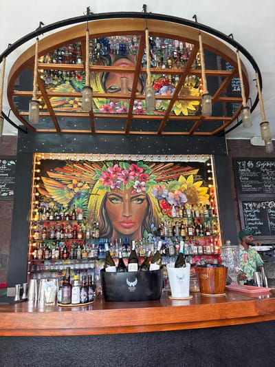 The image captures the interior of a bar, possibly located in Talpe, Sri Lanka, as the bucket says "Angel Beach". A large, circular wooden frame with a dark metal exterior holds a mirror that reflects the bar's back wall, creating a visually interesting doubled effect. The bar is well-stocked with various bottles of alcohol arranged on shelves behind a vibrant mural depicting a woman with floral headwear and a hummingbird. String lights surround the mural. A wooden bar top is visible in the foreground with wine bottles in ice buckets and tools used to make drinks. At the top, a menu board is visible with drink options written in chalk. There is also a person dressed in a green shirt with a hat. The bar's atmosphere is lively, as indicated by the colorful mural, lighting, and assortment of beverages.