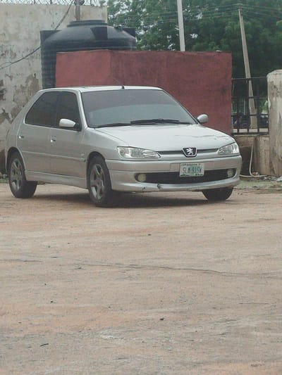 A silver Peugeot 306 is parked on a dirt surface in front of a red wall, a large black water tank, and a white concrete structure. The car has a license plate that reads "SLM-91SW". The background shows some trees and utility poles. The scene appears to be in an outdoor urban environment, possibly a residential area or a less developed part of a city. The overall impression is that of a parked vehicle in a somewhat neglected or rustic setting.