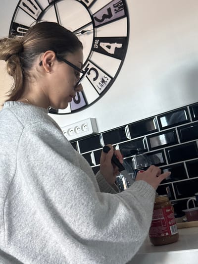 The scene depicts an indoor environment, likely a kitchen, located in Limeil-Brévannes, France. A female individual, shown in profile facing right, is engaged in an activity. She has light-brown hair tied back in a bun, wears black-framed glasses, a small gold hoop earring in her left ear, and a thin gold chain necklace. Her attire consists of a light grey, possibly knitted, long-sleeved top. Her visible left hand, with black fingernail polish, holds a knife with a black handle and a silver blade, positioning it over the opening of a brown-colored jar. Her right hand is also near the jar's opening. The jar features a red label with the word "Creamy" partially visible, suggesting a food product.
The background features a wall divided horizontally. The lower section consists of glossy black rectangular subway tiles. Above these tiles, a white multi-socket power strip with four European-style outlets is mounted. The upper section of the wall is plain white, on which a large, circular wall clock is mounted. This clock has a black frame, a face with alternating white and dark segments, black numerals (e.g., 3, 4, 5, 6, 9, 10, 11, 12 are partially visible), and specific text "LAKES," "1948," and "OHIO." The clock hands indicate approximately 10:50. A clear plastic bottle is visible behind the knife and jar. The objects rest on a light-colored countertop. To the right of the jar, a small tray holds two small cups, one red and one white with a red rim.