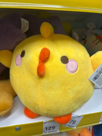 This is a close-up, eye-level photograph of a yellow, plush chicken toy displayed on a store shelf. The toy has a round body, small orange feet, and a small orange beak-like feature. It has two brown eyes and two pink circular cheeks outlined in pink stitching. To the left of the chicken toy, a portion of an orange plush toy is visible, and behind it, a purple plush toy is partially seen. The shelf is white and has a yellow trim along its front edge. A price tag with the number "79" is visible on the shelf in the lower right portion of the image. The background is blurred and shows other merchandise on shelves. No people are present in the image. The image does not provide any information regarding the city of Ivanivka, Ukraine.