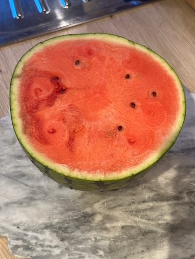 A half watermelon rests on a marble countertop in a kitchen. The watermelon is bright red with some visible seeds and a green rind. In the background, a stainless steel appliance with blue glowing elements is visible, suggesting it could be a gas stove or grill. The countertop has a marbled pattern. The scene appears to be indoors, likely in a residential kitchen. There is no indication of this location being in the city of Kiel, Germany.