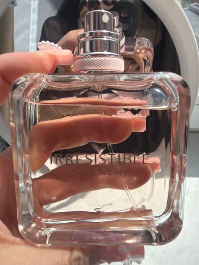 A close-up, brightly lit image features a right hand holding a clear glass perfume bottle containing a pink-tinted liquid, approximately three-quarters full. The bottle's front is embossed with the word "IRRESISTIBLE" in prominent dark, reflective capital letters, with "GIVENCHY" inscribed below it in a smaller font. The top of the bottle displays a metallic rose gold and silver sprayer mechanism, detailed with a textured, light pink band below the cap and a clear plastic dip tube extending into the fragrance. The hand exhibits manicured nails with a clear, iridescent beaded embellishment on the index finger. A thin gold-colored ring is visible on another finger. In the highly reflective background, partially seen within a white-framed mirror, is the reflection of a person wearing dark, possibly patterned clothing, holding a dark electronic device, likely the camera used for the photograph. Strong, directional lighting, possibly natural sunlight, creates numerous highlights and reflections across the bottle's surface. The location is in the city of Novosaratovka, Russia.