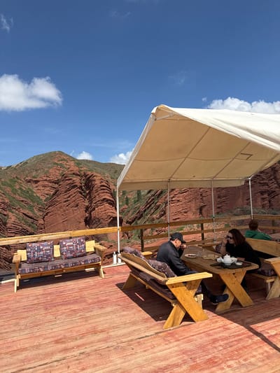 This image captures an outdoor dining or relaxation area on a raised wooden deck, overlooking the distinctive red rock formations of Jeti-Ögüz, Kyrgyzstan. A large, cream-colored canopy provides shade over parts of the deck.
Two main individuals are seated at a rustic wooden table with benches. A man, wearing a black cap and black top, is on the right, looking down. Opposite him, a woman with long dark hair, wearing a black top and sunglasses, is seated. On their table, a white teapot, cups, and small plates with food are visible, suggesting they are enjoying a meal or drinks. A third person is partially visible behind the woman. The deck is furnished with multiple sturdy wooden tables and benches, some featuring colorful, patterned cushions.
Beyond the outdoor seating area, the background is dominated by the dramatic red sandstone mountains, characteristic of the Jeti-Ögüz region. These formations display prominent horizontal layering and are sparsely covered with patches of green vegetation. The sky above is a vibrant, clear blue with a few scattered white cumulus clouds, indicating bright daylight and good weather. The scene conveys a relaxed atmosphere, offering a scenic spot for visitors to enjoy the natural beauty. No discernible text is visible within the image.
