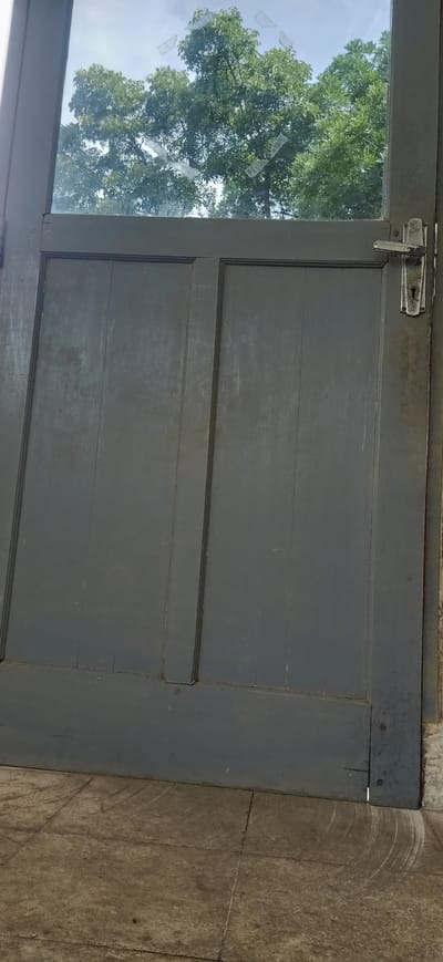 The image shows a weathered, dark gray wooden door with two panels. The top panel of the door is a glass pane, which is partially cracked and has several pieces of tape over the cracks. Through the glass, lush green trees and a cloudy sky are visible, suggesting it is daytime. On the right side of the door, a silver door handle and a keyhole are present. The door is set in what appears to be an outdoor setting, with a textured, light-colored tiled floor visible at the bottom. The tiles show scuff marks, indicating wear and tear. The door itself has visible wear, with paint peeling in some areas and cracks in the wood. The overall impression is of an older, slightly neglected structure. There is no visible text in the image.