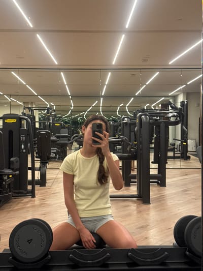 A young woman with braided hair is reflected in a gym mirror while taking a selfie with her phone. She wears a pale yellow t-shirt and grey athletic shorts. She is standing at a dumbbell rack holding a medicine ball between her thighs. Behind her, the gym is equipped with rows of weight machines and bright overhead lighting. The floor appears to be a light wood-tone.