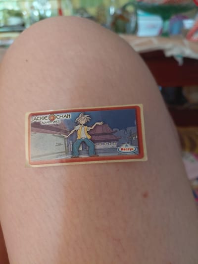 The image shows a close-up of a sticker that features artwork from the animated television series "Jackie Chan Adventures." The sticker is placed on a person's skin, likely an arm or leg, as evidenced by the texture and natural color variations. The artwork depicts Jackie Chan in a dynamic pose, appearing to be mid-action. He is illustrated in his characteristic attire: a yellow vest over a white shirt and blue pants. The background of the sticker suggests an urban or traditional Asian architectural setting, with buildings rendered in a cartoon style. A red border frames the artwork, and a small oval logo with the word "Hentai" is visible in the upper right corner of the sticker. Below the illustration of Jackie Chan, text clearly reads "JACKIE CHAN ADVENTURES." The lighting in the image suggests it was taken indoors during the daytime, with no specific indication of the weather or precise location beyond the provided context of Marynivka, Ukraine. The overall presentation implies a casual setting where the sticker is being displayed.