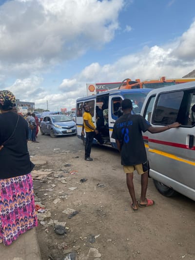 The scene depicts a busy unpaved roadside area in Ablekuma, Ghana, under a partly cloudy sky. The foreground consists of compacted dirt heavily strewn with debris, including cardboard, plastic, and dark refuse.

On the far left, a person, viewed from behind, wears a black top, a colorful patterned skirt (pink, purple, orange, black geometric design), and a dark head covering with metallic accents. Adjacent to this individual, a paved road features a light blue Toyota Yaris hatchback (license plate GH-540-20 visible) stopped in traffic, with several indistinct individuals standing nearby.

In the midground, a blue and white passenger minibus (trotro) is parked on the dirt shoulder with its side door open. A man in a yellow t-shirt and dark trousers stands next to the open door, smiling and looking towards the camera. To his right, a young male, viewed from the back, wears a black t-shirt with "STAFFORD JR 8" printed in blue, light brown shorts, and mismatched slides (one pink, one dark). He leans on the open window frame of a silver passenger van parked directly to the right of the minibus.

The background features commercial structures, including a prominent red and yellow Shell gas station sign and a red and white KFC restaurant sign. An orange tanker truck is partially visible behind the blue minibus. The overall environment suggests a bustling urban transport hub.