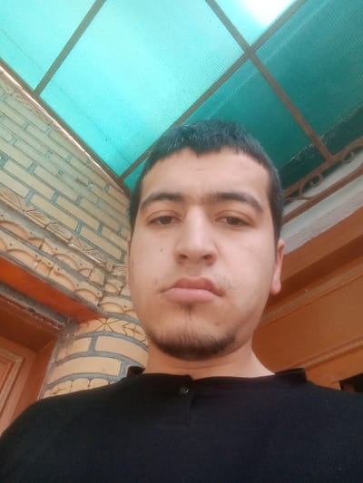 This image, captured in Kasan, Uzbekistan, depicts a male subject positioned centrally, directly facing the camera. He has short dark hair, dark eyes, connected eyebrows, and a visible shadow of stubble or a short beard on his chin and upper lip, maintaining a neutral expression. He is wearing a dark-colored collared shirt. The background features a light-colored brick wall with decorative relief patterns on the left, partially revealing a dark wooden door or frame. To the right, a darker wooden trim and an ornate dark metal scroll are visible. Overhead, a translucent turquoise corrugated roof panel, supported by a dark grid frame, covers the area, indicating a covered exterior or semi-enclosed space. The scene is illuminated by diffused, ambient light.