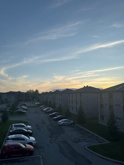 The image depicts a sunset over a residential area in Gatineau, Canada. The sky is a soft blue with wispy clouds, illuminated by the warm glow of the setting sun on the horizon. In the foreground, a parking lot is filled with various cars, including a red SUV, a white SUV, and several sedans, parked in designated spots. Adjacent to the parking lot are rows of modern apartment buildings constructed with grey brick. Small, dark green evergreen trees are scattered throughout the scene, lining the buildings and the parking lot. The overall atmosphere is one of tranquility and domesticity as daylight fades.