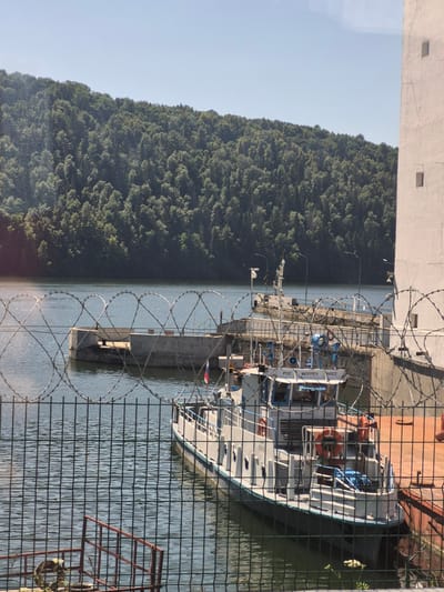 This image shows a view of a body of water in Pavlovka, Russia. In the foreground, there is a metal fence with barbed wire. Beyond the fence, a boat is docked near a concrete structure. The boat is white with blue trim and has Russian text visible on its side. In the background, there is a densely wooded hillside under a clear blue sky. To the right of the scene, a tall, light-colored building with small square windows is partially visible. The water is calm, reflecting the surrounding greenery.