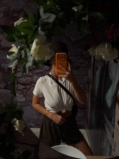 A woman in a white knotted t-shirt, dark shorts, and a dark hat takes a mirror selfie in a bathroom. She holds an orange smartphone to obscure her face. The bathroom features a stone-textured wall and a distressed wooden door. In the foreground, a mirror reflects the woman and the surroundings, with artificial white roses and greenery arranged around its edge. A white sink is partially visible in the lower right corner of the reflection. The lighting is dim and moody. The location is identified as Urbanització Cales I Talaies, Spain.