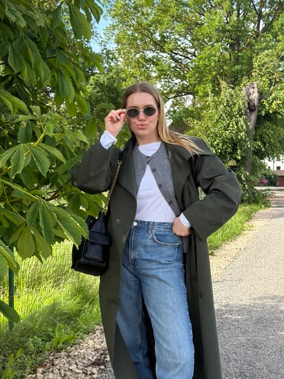 In this vertical outdoor shot, a young woman with blonde hair and sunglasses is posed against a backdrop of lush green trees and a gravel path. She is wearing a white t-shirt layered under a grey cardigan and an olive-green trench coat. Her blue jeans are visible, and she is holding her sunglasses, which are round and black-rimmed, with her right hand. A small black quilted bag with a gold chain strap is visible at her hip. The scene suggests a casual, everyday moment, possibly a walk outdoors. The lighting indicates it is daytime, and the abundant greenery suggests a warm season, likely spring or summer. The location context provided is Dreiliņi, Latvia, which aligns with the natural, outdoor setting depicted. No text is visible in the image.