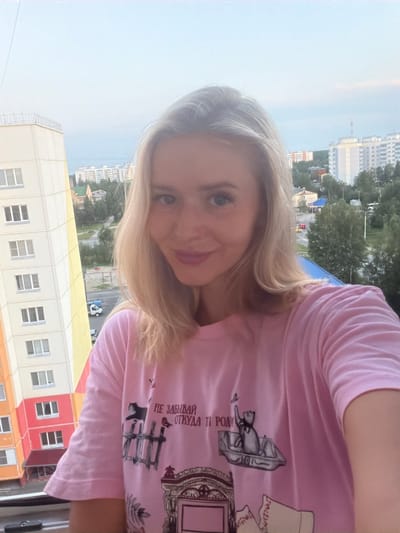 A blonde woman is taking a selfie on a balcony in Pyt'-Yakh, Russia. She is wearing a pink t-shirt with a graphic print. In the background, there are residential buildings, trees, and a street with a white truck. The sky is overcast.
