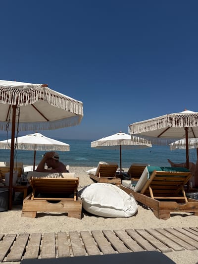 The image depicts a beach scene with a clear blue sky and calm blue water. Several white beach umbrellas with decorative fringe are set up on the sand. In the foreground, a wooden lounge chair with a green cushion is visible, along with a white beanbag. A person wearing a white cap is lying on a lounge chair in the background, with their hands clasped behind their head. The lighting suggests it is daytime, likely late morning or early afternoon, with the sun casting shadows on the sand and wooden decking. The setting appears to be a beach resort or a private beach area in Pallini Municipal Unit, Greece, given the context. The overall mood seems relaxed and tranquil.