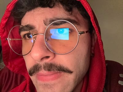 The upside-down image features a person wearing round glasses and a red hooded sweatshirt. One lens reflects a blue light, possibly from a screen. The person has dark eyebrows and a light complexion. The background is a neutral wall. The composition is a close-up of the face, emphasizing the glasses and the reflection.