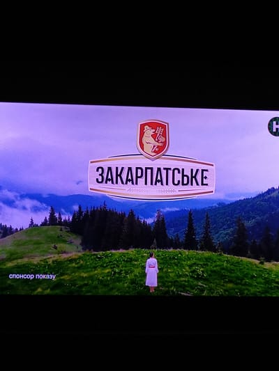 The image depicts a vertical view of a scenic mountain landscape with a logo and text. In the foreground, a white sign with a distinctive red and yellow shield logo is prominently displayed. The shield contains imagery of a bear and tools, possibly representing local heritage or industry. The sign itself has bold, vertical text in a foreign language, appearing to read "ЗAKAPПАТЬСЬКЕ" (Zakarpatske). Behind the sign, a woman in a white dress stands in a lush green meadow on a hillside. The background features a dense forest of dark green pine trees cascading down a mountain slope, with a hazy, purple-tinged sky and distant mountains visible. The scene suggests a natural, possibly rural or mountainous, setting. The lighting and muted colors indicate it could be late afternoon or early morning, with possibly overcast or misty weather. Towards the bottom right of the image, small, light-colored text reads "спонсор показу" which translates to "sponsor of the show" in Ukrainian.