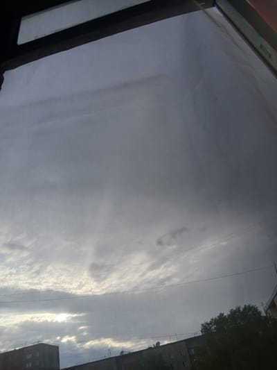The image shows a view through a window screen of a cloudy sky over a city. The sky is mostly overcast with some breaks where the sun is visible, creating bright patches. In the foreground, the window screen is clearly visible, creating a grid pattern over the scene. Below the sky, parts of buildings and trees can be seen. The buildings appear to be apartment blocks, with multiple windows and some visible rooftop structures. The overall impression is a somewhat gloomy, overcast day, possibly late afternoon or early morning, as indicated by the low angle of the sun peeking through the clouds. The location is identified as Aksu, Kazakhstan. There are no people or specific actions depicted in the image.