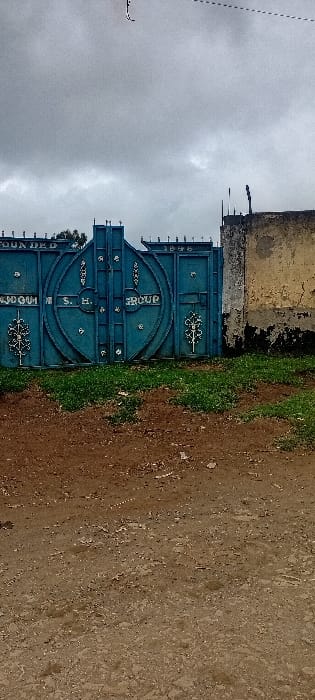 A blue metal gate stands against a weathered, yellow-toned wall. The gate has decorative flourishes and the words "FOUNDED 1970", "S.H. GROUP" displayed on it. Overcast sky above. Dirt path with vegetation is on the right side.