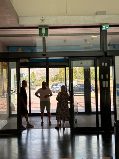 The image shows the interior of a modern building, likely a commercial establishment, with large glass doors and windows. The scene is viewed from inside, looking outwards through the doors. Sunlight streams in, casting long shadows on the polished floor, suggesting it is daytime, possibly late morning or afternoon. The weather appears to be sunny and warm, as evidenced by the bright light and the casual attire of the people.
In the foreground, three people are visible, silhouetted against the bright light from outside. A woman in a patterned dress stands near a set of glass doors. Further inside, a man in a light-colored polo shirt and shorts is seen. Nearer to the camera, another man in a dark t-shirt and shorts is seated. Their postures suggest they are either entering or exiting the building, or perhaps waiting.
Through the glass, an outdoor scene is visible, with a street or pathway and some greenery. Cars are parked or driving in the background, and a shopping cart is also visible. The architecture of the building appears contemporary, with dark framing around the glass panels.
A prominent green and white sign with an arrow and a figure is visible on the left side of the image, indicating an exit. The text on this sign is in Italian and likely refers to an emergency exit. There may be other signs with text visible on the doors or windows, but they are largely obscured by reflections or the silhouettes of the people. The overall impression is one of everyday activity in a well-lit, modern commercial space.