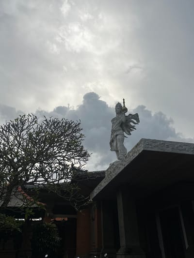 The image shows a cloudy sky above a building in Celuk, Indonesia. A stone statue of a person with ornate wings and an instrument is fixed atop the building's edge. A tree with a dense network of branches is visible below. The building is made of dark wood, with intricate carvings along the edge.