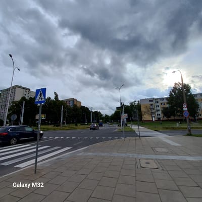 The image depicts an urban street scene in Olsztyn, Poland, on an overcast day. In the foreground, a pedestrian crossing with white diagonal stripes is visible on the asphalt road, bordered by a sidewalk paved with grey square tiles. A dark blue SUV is parked to the left, partially obscuring the pedestrian crossing. A signpost with a blue square sign displaying a white pedestrian silhouette is planted on the sidewalk. Beyond the road, several apartment buildings with multiple stories and numerous windows are situated, interspersed with a dense cluster of green trees. Streetlights with curved arms extend over the roadway. In the distance, other vehicles are visible on the road, and more buildings and trees populate the landscape. The sky is filled with heavy, grey clouds, with a sliver of bright sky peeking through on the right side, suggesting the sun might be present behind the clouds. The "Galaxy M32" watermark is present in the bottom left corner of the image.