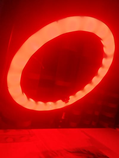 The image shows a red ring light, which is a type of lighting equipment often used for photography and videography. The ring light is illuminated with a warm, orangish-red light, casting a strong red glow on its surroundings. The background appears to be a dark, possibly textured surface, with some subtle vertical lines suggesting curtains or a backdrop. The lower portion of the image shows a flat, reflective surface also illuminated by the red light, with some indistinct patterns or creases visible. The ring light is suspended or mounted on a stand, the base of which is partially visible at the bottom of the frame. There are no people or animals present in the image. The overall impression is one of artificial lighting used to create a specific mood or effect. There is no information in the image that would allow for identification of its location in Markovo, Bulgaria.