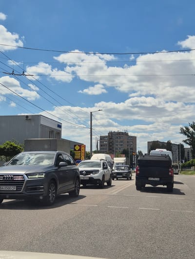 The image depicts a street scene in Kropyvnytskyi, Ukraine, on a sunny day with scattered clouds. Traffic is moderate, with several vehicles visible on the asphalt road. In the foreground, a dark grey Audi Q7 SUV is stationary, its license plate reading "BA 0500 CP". Behind it, a white Renault Duster SUV is also stopped, with its license plate "AI 7851 OO". Further ahead, a dark-colored pickup truck with a black cover over the bed is in motion, its license plate "BA 6782 BA". Other vehicles, including vans and a older model sedan, are also present in the traffic flow.
The background reveals a mix of urban and commercial structures. On the left, a modern, grey building with windows and an air conditioning unit is visible. A sign with text, possibly advertising furniture, is also present. Beyond that, taller, multi-story apartment buildings dominate the skyline. Trees line the side of the road, and power lines are strung overhead, crossing the blue sky dotted with white clouds. The overall impression is a typical urban environment experiencing light traffic under a clear sky.