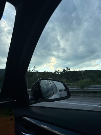 An exterior, eye-level, wide shot shows a view from inside a vehicle looking out the side window and through the side mirror. The sky is cloudy with patches of sunlight breaking through. The landscape outside includes a forested hillside and a highway. A side-view mirror reflects the car's interior and the sky. The location is identified as Dolno Varshilo, Bulgaria, though no specific landmarks are visible to confirm this. The scene depicts travel on a road, with the environment suggesting a rural or semi-rural setting under a dynamic sky.