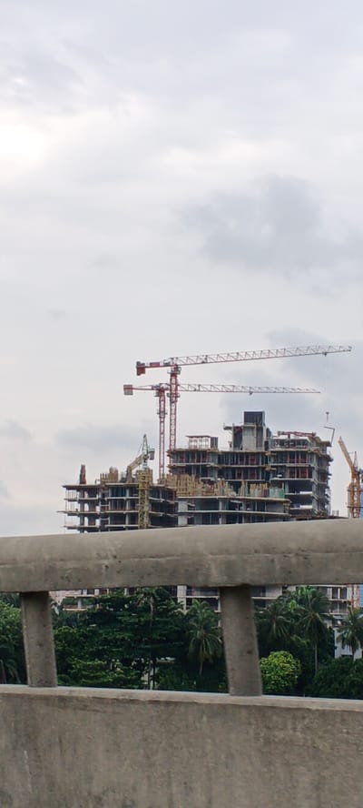 In Ikoyi, Nigeria, a massive construction project is underway, dominated by a partially built, multi-story structure. Several construction cranes are visible, including two prominent red tower cranes reaching high into the overcast sky, positioned to the left and center of the frame. A yellow tower crane is also present, situated to the left of the main building. The building itself is a skeletal framework of concrete and steel, with visible floors, columns, and scaffolding. Below the construction site, lush green trees and palm trees create a dense canopy, suggesting a tropical environment. In the foreground, a concrete barrier, possibly part of a bridge or elevated walkway, partially obstructs the view. The scene is devoid of any people. The lighting indicates an overcast day.