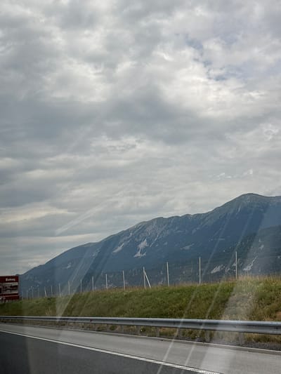 The image shows a view from a vehicle driving on a road. In the foreground, there is a paved road with a metal guardrail. Beyond the road, there is a grassy embankment with a fence line. In the background, there are mountains covered in vegetation and some patches of snow. The sky is overcast with clouds. On the left side of the image, there is a brown road sign with text and images, appearing to indicate a destination or landmark. The sign reads "Bohinj" at the top. The location is Radovljica, Slovenia.