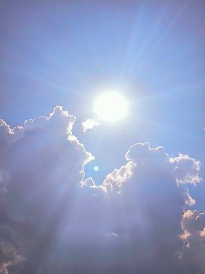 The image shows a bright sun with visible rays shining through clouds against a clear blue sky. The clouds are a mix of white and gray, with some areas illuminated by the sun. The overall impression is of a sunny day with scattered clouds. There is no information in the image to suggest the location is in Vētras, Latvia, or any specific city. The scene is entirely natural, depicting the sky and sun. There are no people, subjects, objects, or actions depicted.