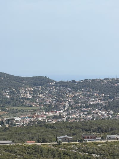 This panoramic photograph, captured in Olèrdola, Spain, showcases a vast residential landscape built into and around wooded hills under a clear, bright sky. The upper third of the image is dominated by a pale blue sky, indicating serene, sunny weather, likely midday or early afternoon.
Below the sky, a distant, undulating ridge line, thickly covered in dark green trees, forms the horizon. As the land descends towards the foreground, the hillsides become more extensively populated. Numerous houses, featuring light-colored facades and reddish-brown tiled roofs, are densely clustered, forming a sprawling town or village that blends with the natural contours of the terrain. The vegetation primarily consists of dense, evergreen trees, characteristic of a Mediterranean climate.
Further into the foreground, a prominent band of forest stretches horizontally, acting as a natural divider. Across this forested section, several parallel power lines are visible. Beyond this, to the right, a series of more contemporary buildings with distinct architectural styles are sparsely arranged along what appears to be a road or path, with further dense vegetation on a steeper rise beyond them. The overall scene depicts a harmonious, yet extensive, integration of urban development within a hilly, wooded environment. No human activity or visible text is present.