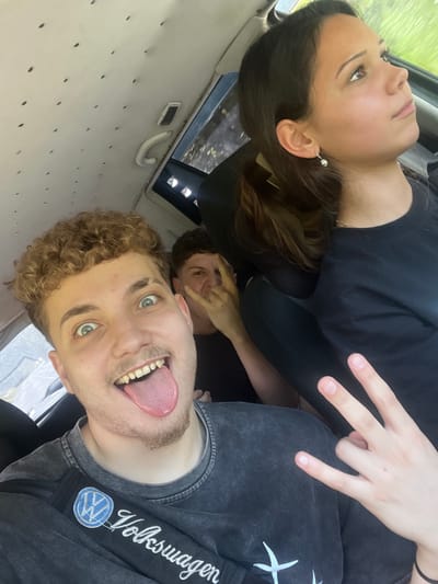 Three individuals are inside a vehicle. The person in the foreground, a male with curly blonde hair, is sticking his tongue out at the camera with his eyes wide open. He is wearing a dark grey t-shirt with a "Volkswagen" logo on a strap across his chest. To his left, partially obscured, is another male with short brown hair, making a peace sign with his fingers and a surprised expression. On the right, a female with dark hair pulled back in a ponytail looks up and to the side, wearing a dark t-shirt and a small earring. The interior of the vehicle appears to be a van, with a textured, cream-colored ceiling with small dark circular indentations. A white grab handle is visible near the window. The lighting suggests it is daytime, possibly overcast, with some light filtering in from the windows. The backdrop outside the windows is blurred, but appears to be greenery, suggesting a rural or suburban setting. There are no specific landmarks or indications that definitively place the location in Vlahovo, Bulgaria, but the overall scene is consistent with being in a vehicle in a community.