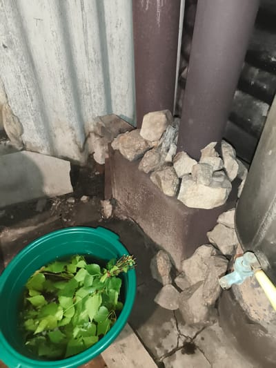 In Il'mino, Russia, a green plastic basin filled with water and birch branches is positioned on the ground next to a corrugated metal wall and two large, dark metal pipes. The pipes are surrounded by a collection of light-colored stones. A light blue faucet with a yellow hose attached is visible to the right of the pipes. The ground appears to be concrete and dirt, with some debris scattered around. The scene suggests an outdoor or semi-outdoor setting, possibly a utility area or bathhouse.