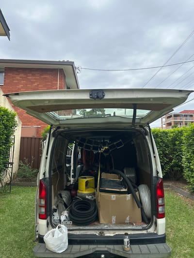 The back of a white van is open, revealing a cluttered interior filled with various tools and equipment. A cardboard box, rolls of wire, black tubing, a yellow container, and metal framework are visible. The van is parked on a grassy area with a house and greenery in the background. The van's open door frames the scene, highlighting the disarray within.
