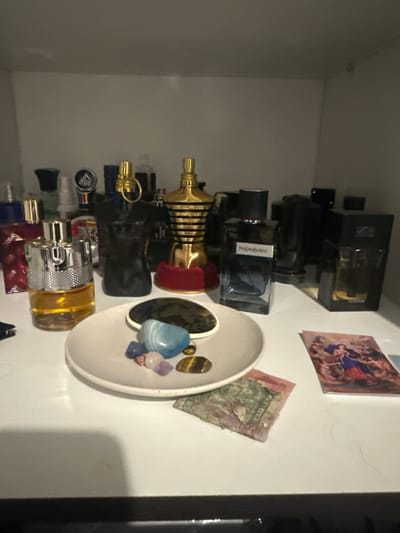 A collection of perfume bottles is arranged on a shelf, ranging from black square bottles to a gold, torso-shaped one. A white dish holds a cluster of colorful stones, including blue agate, tiger's eye, and amethyst. A dollar bill and a small art print are affixed to the wall beside the shelf.