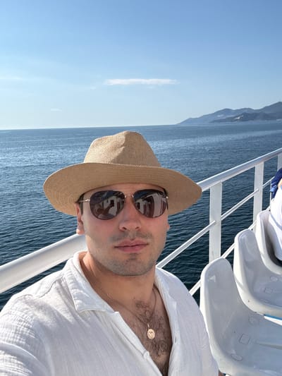 A man wearing a straw hat and sunglasses is taking a selfie on a ferry. He is dressed in a white shirt and is wearing a necklace. The background shows the sea and a coastline with mountains. The seats of the ferry are visible on the right side of the image. The location is on the sea, near Thassos, Greece.