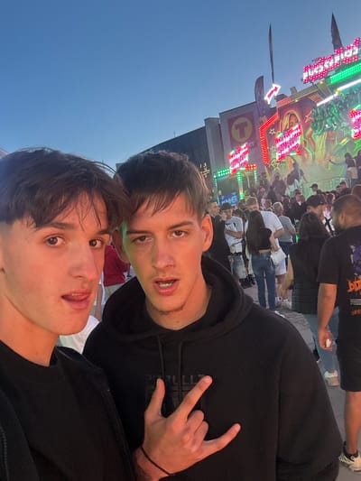 Two young men pose for a selfie at a fairground. The man in the foreground wears a black shirt, while the other, slightly behind, dons a black hooded sweatshirt and flashes a "rock on" hand gesture. Behind them is a crowd of people and a lit-up carnival ride, and a blue sky is visible in the background. The location may be Bozhurishte, Bulgaria.