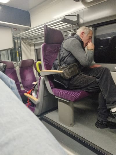 An older man sits in a purple train seat, dressed in a gray sweater, black vest, and black pants. He has a small black bag slung across his body and appears to be coughing into his hand. The train interior is visible with overhead luggage racks and rows of purple seats. The window shows a dark, unclear view of the outside. Another person's jeans-clad leg is partially visible in the foreground.