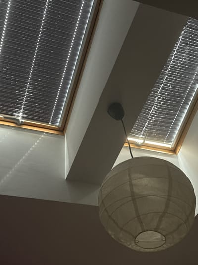 The image shows a view looking up towards two angled skylights with blinds. A white, spherical paper lantern hangs from a black cord. The skylights are framed with wood. Light is filtering through the blinds, creating a pattern of bright lines on the white ceiling below. The environment appears to be an interior space, possibly a room or attic, with a sloped ceiling. The location is specified as Poznan, Poland. There are no people or discernible actions depicted.