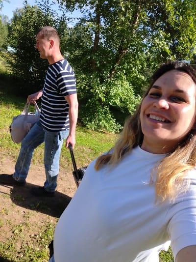A man and a woman are outdoors on a sunny day, with green trees and foliage in the background. The man, in profile to the left, wears a striped t-shirt and jeans. He is carrying a grey duffel bag in his left hand and pulling a black rolling suitcase with his right hand. The woman is in the foreground, taking a selfie. She is smiling broadly and wearing a white t-shirt. Her hair is light brown and falls around her shoulders. The setting appears to be a park or natural area, with a dirt path and grass visible. The time of day is likely daytime due to the strong sunlight and shadows. The overall mood is cheerful and relaxed, suggesting a casual outing or trip. No text is visible in the image.