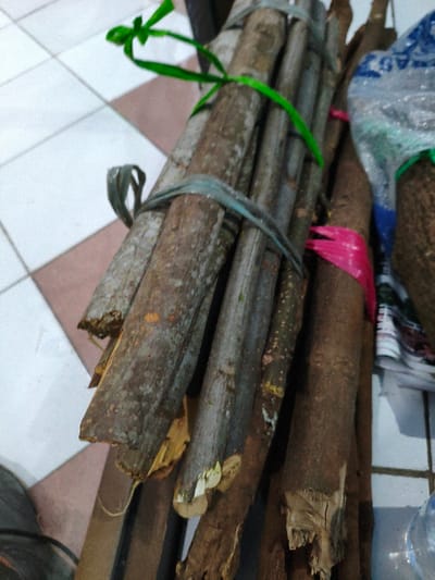 A close-up, slightly high-angle view depicts several bundles of wooden sticks or branches resting on a tiled floor. The floor consists of white and light brown/beige square tiles arranged in a diamond pattern. The wooden pieces vary in diameter and bark texture, with some ends exhibiting fresh breaks or cuts.
One prominent bundle, located centrally, is secured by two light blue plastic straps. Above this, another bundle or section of wood is tied with a bright green plastic strap. To the right, a darker bundle of wood is secured with a bright pink plastic strap. Behind these bundles, a partially obscured clear plastic bag, possibly containing items, is visible, along with what appears to be a stack of papers or magazines. The lower right foreground shows a portion of a transparent plastic bottle. The lower left foreground reveals a dark, indistinct object, possibly part of a tire or a dark hose. The scene takes place indoors, in the city of Muka, Indonesia. No individuals are present.