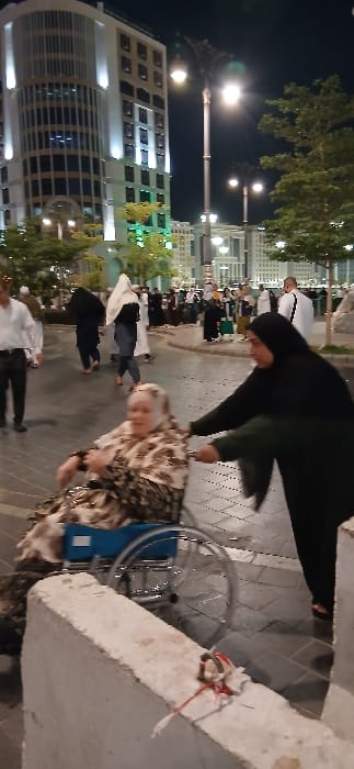 The image depicts a nighttime urban scene in a pedestrian area near Al-Masjid an-Nabawi in Medina, Saudi Arabia. In the midground, a woman dressed in a black abaya and hijab pushes an older woman seated in a blue and silver manual wheelchair. The older woman wears a patterned headscarf and garment. Her expression is indistinct. The pusher's right foot, clad in a sandal, is visible on the tiled pavement. In the foreground, a light-colored concrete barrier extends across the bottom, with a small object wrapped in red and white string resting on its surface. The background features numerous pedestrians, many in Islamic attire, walking across the paved area. Dominating the left background is a tall, modern building with a curved facade and vertical illumination, characteristic of hotels in the vicinity. Ornate black streetlights with bright globes provide overhead lighting, and green foliage from trees is visible on both sides of the walkway. Additional illuminated multi-story buildings are discernible in the distance.
